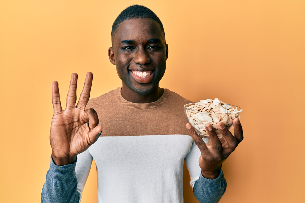 African-American man holding bowl of pumpkin seeds giving the OK sign.